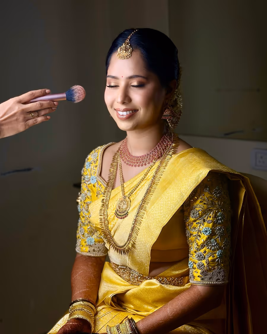 A brush hovering over a bride's cheek in a yellow saree as she smiles